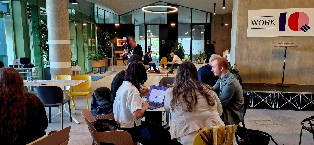 Five people sitting at a round table in a modern collaborative working space. A sign reading 'Work IQ' is hanging on the wall above a stage area.